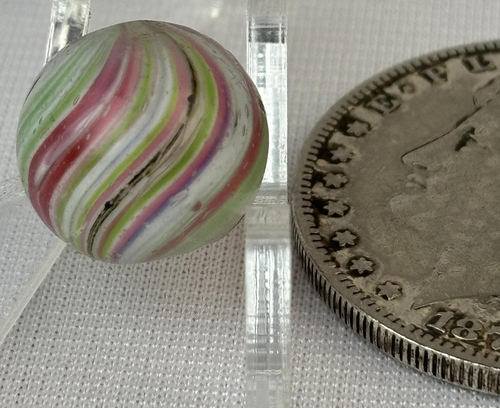 Close-up of a glass marble with concentric colorful swirls—green, pink, red, and white—shown next to a coin for size.