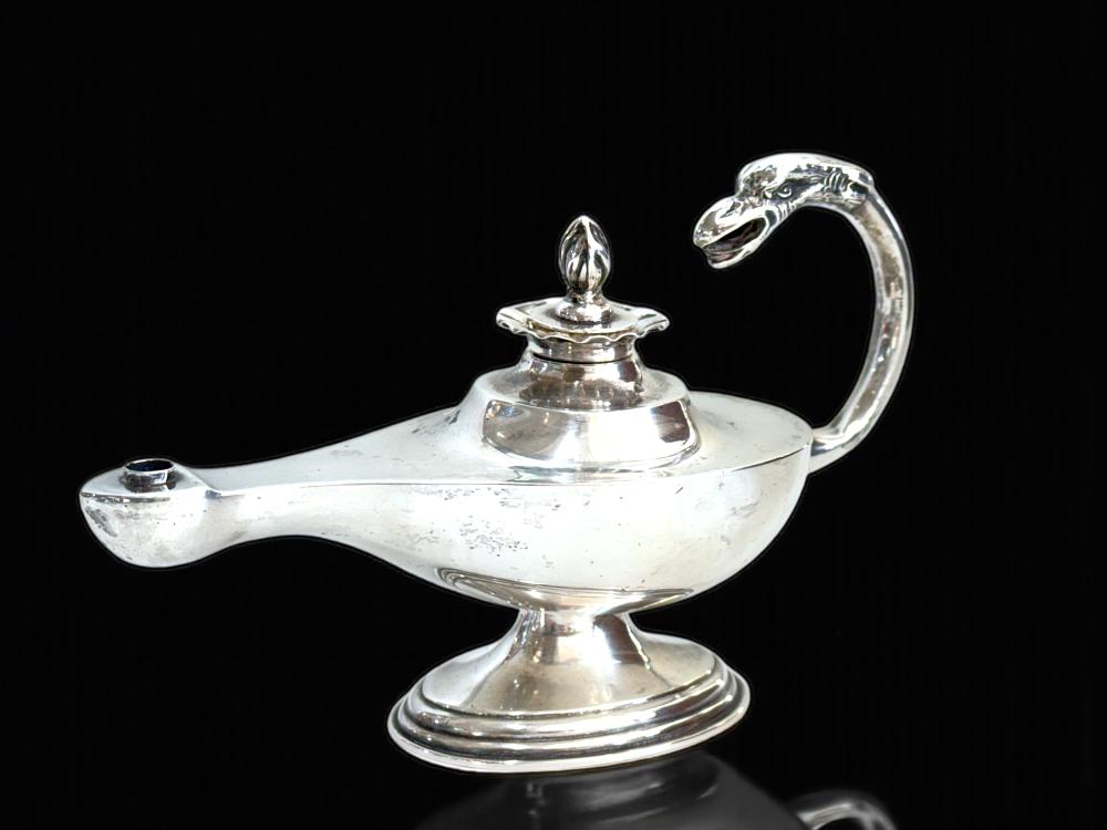 Close-up of a polished silver desk lamp-shaped taper holder with a hinged wick top and curved handle, photographed on a dark background.