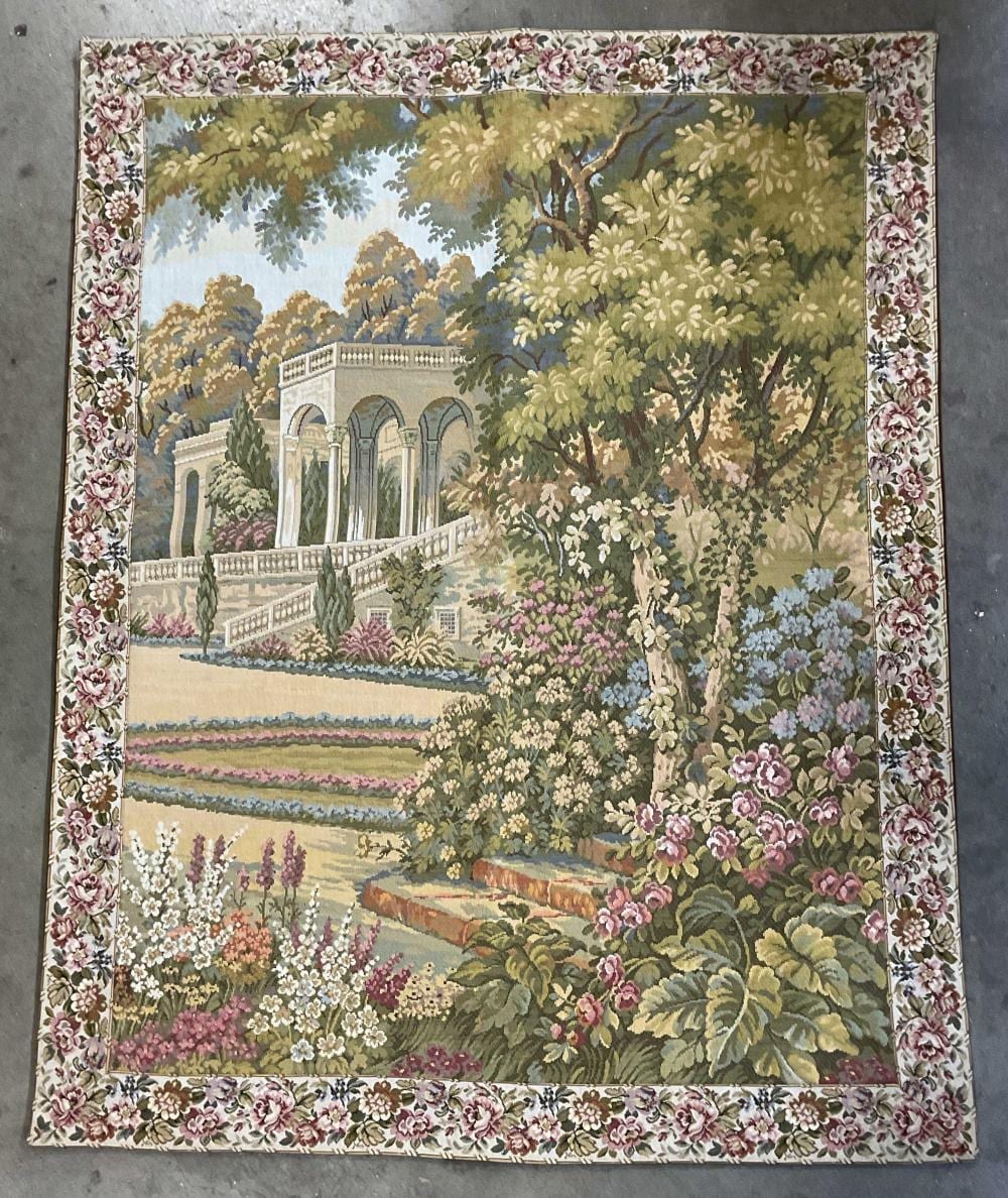 Rectangular needlepoint tapestry showing a garden scene with blooming flowers, layered fields, and an arched building in the background.