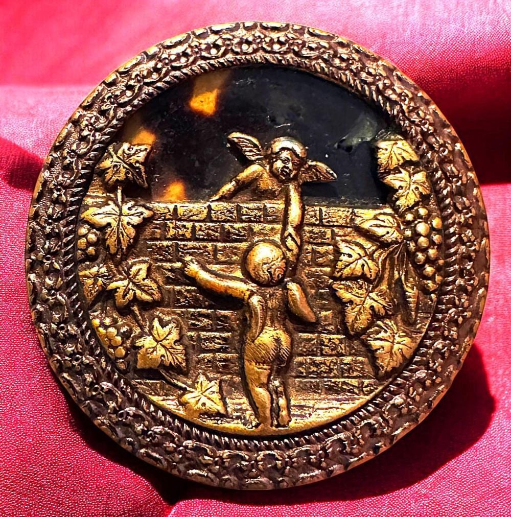 Round dark button with an embossed gold-toned scene of two figures and brick-like steps, shown on a pink fabric background.