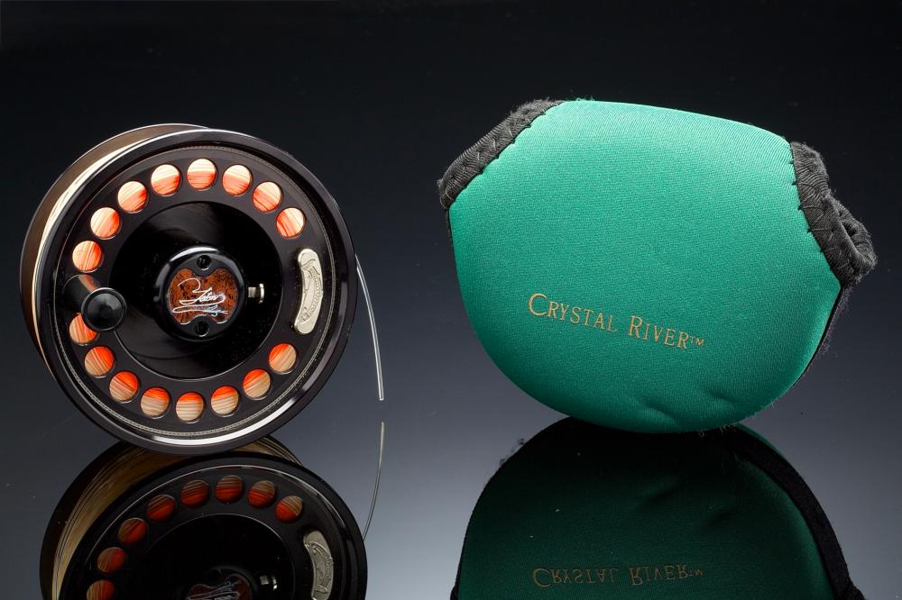 Black fly fishing reel with orange ring of line guide holes beside a green fabric pouch labeled “Crystal Riverm”.