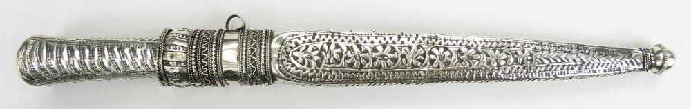 Close-up of a metal dagger with ornate engraved handle and a decorated scabbard, laid horizontally against a white background.