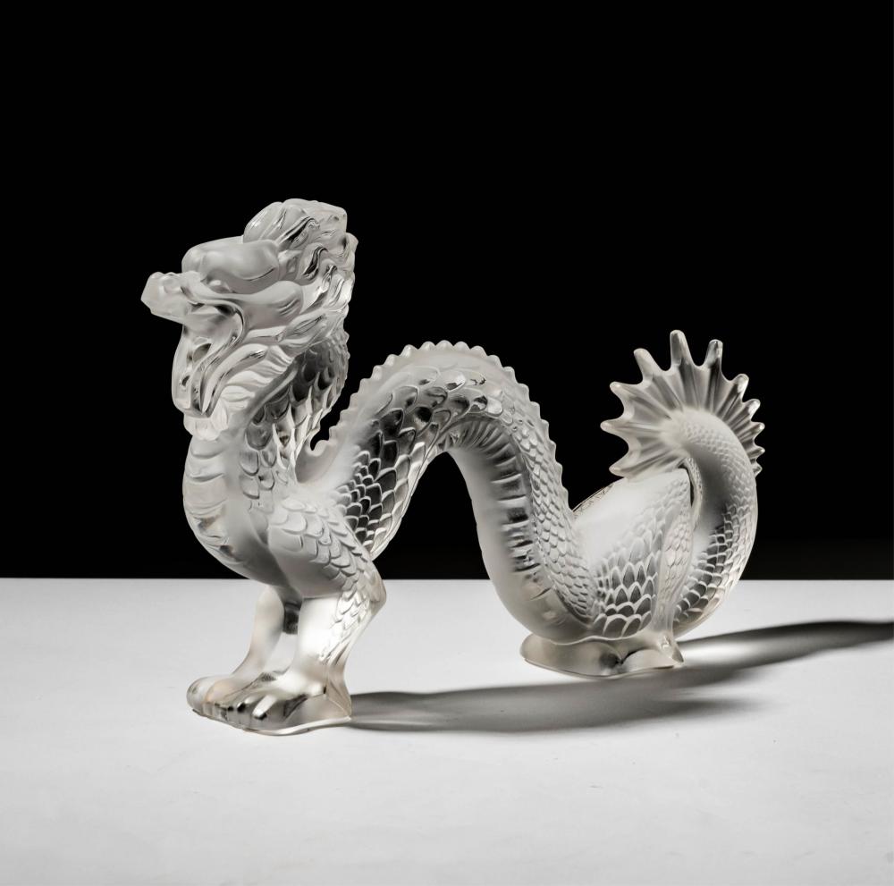 Clear glass dragon figurine with an arched body, detailed scales, and open mouth, photographed on a light surface against a dark background.