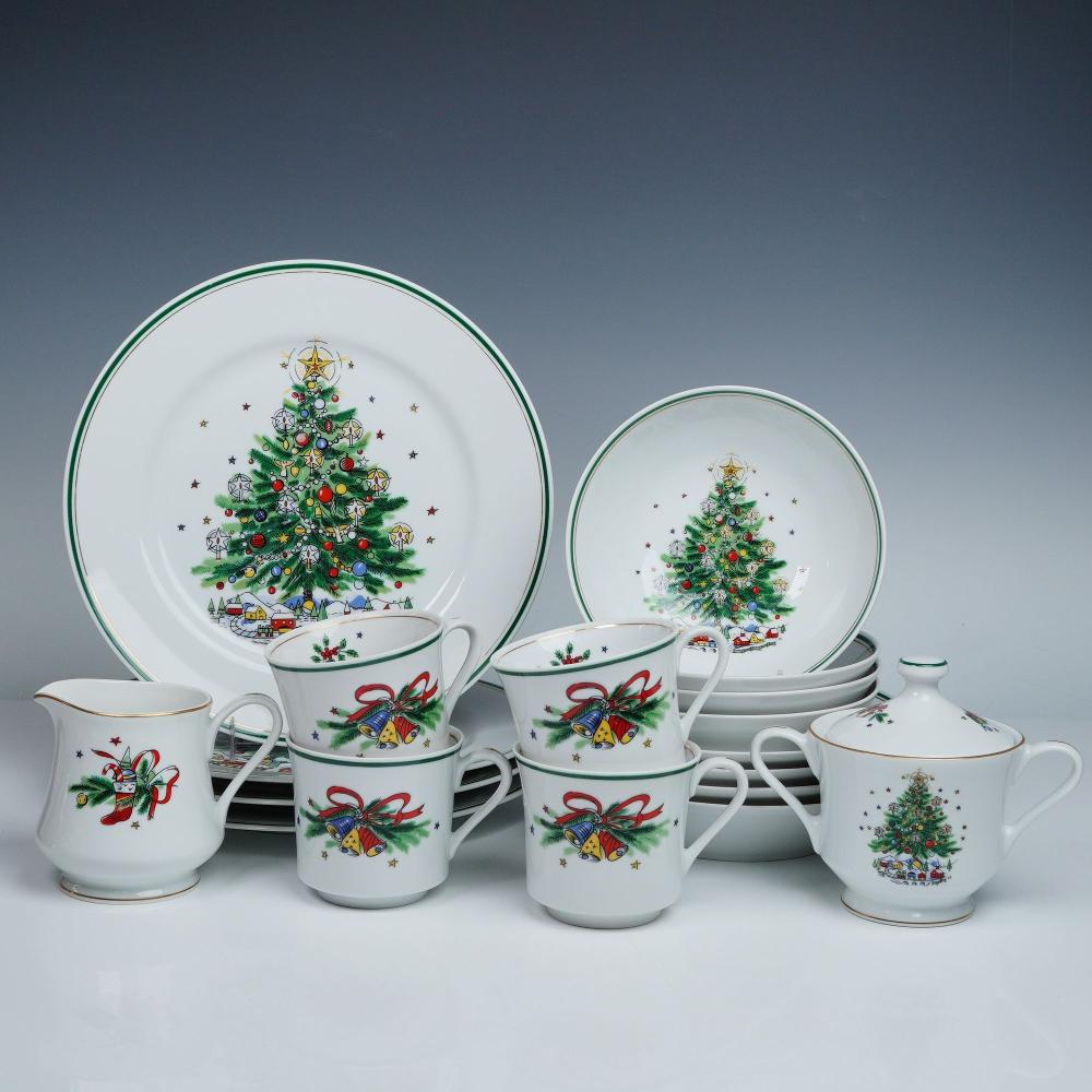 Set of Christmas-themed porcelain dinnerware with plates, teacups, mugs, and a covered creamer featuring a decorated tree and holiday gifts.
