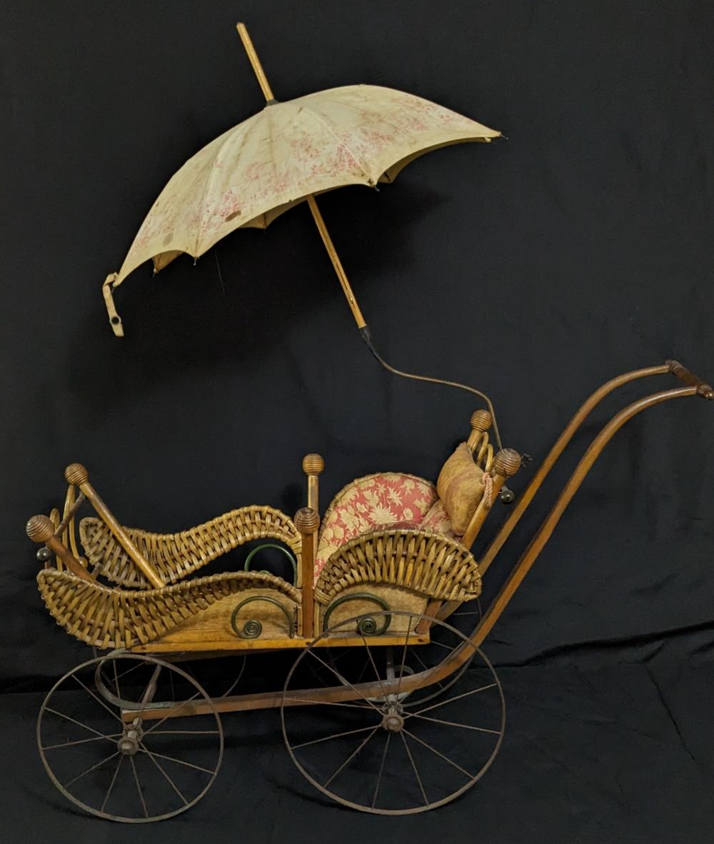 Small wicker doll buggy with curved wheels and a light fabric parasol, set on a dark background.