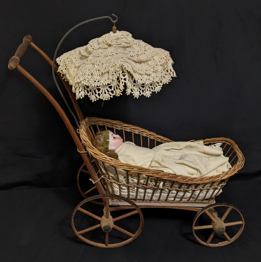 Victorian-style wooden doll carriage with a straw basket body, small doll inside, and an embroidered lace canopy on a curved frame.