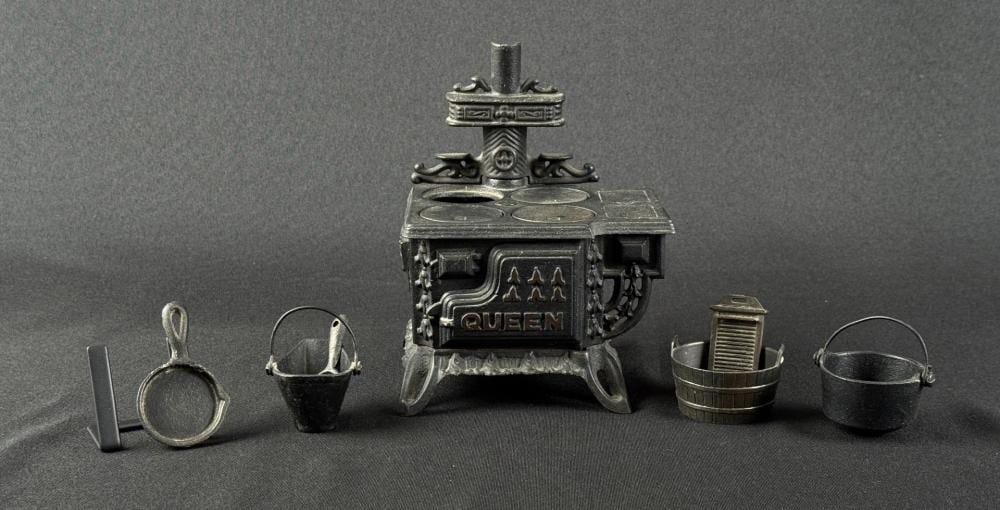 Small cast-iron toy stove marked “QUEEN” with chimney and decorative details, shown with accessories including a pail and a bucket on a dark background.