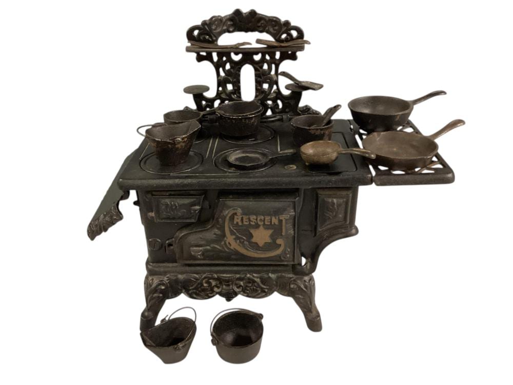 Small cast iron toy stove with decorative base and tabletop accessories, including pots and pans arranged around the burner area.