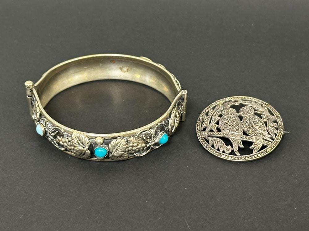 Silver bangle bracelet with floral engraved pattern and blue turquoise-like stones, shown next to a round filigree bird brooch on a dark background.