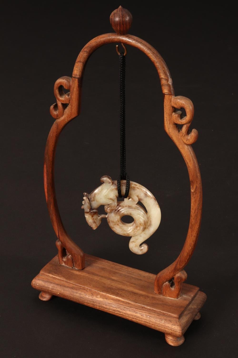 Carved dragon-shaped jade pendant with a dark beaded cord, mounted on a carved wooden stand against a dark background.