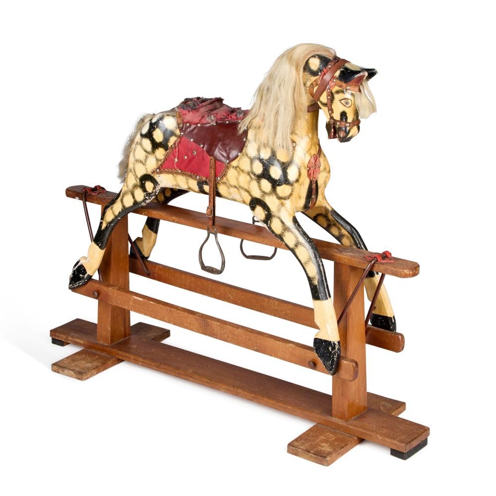 Vintage carved painted wooden rocking horse with cream-and-black spotted coat, red saddle, and long mane on a brown wooden rocker base.