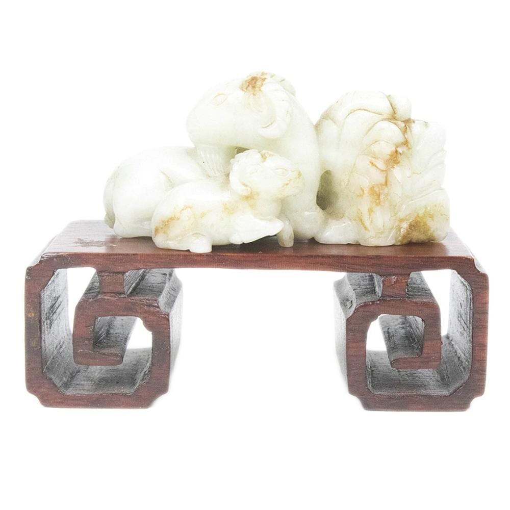 White jade-like carving of three ram figures clustered together on a dark wooden stand.