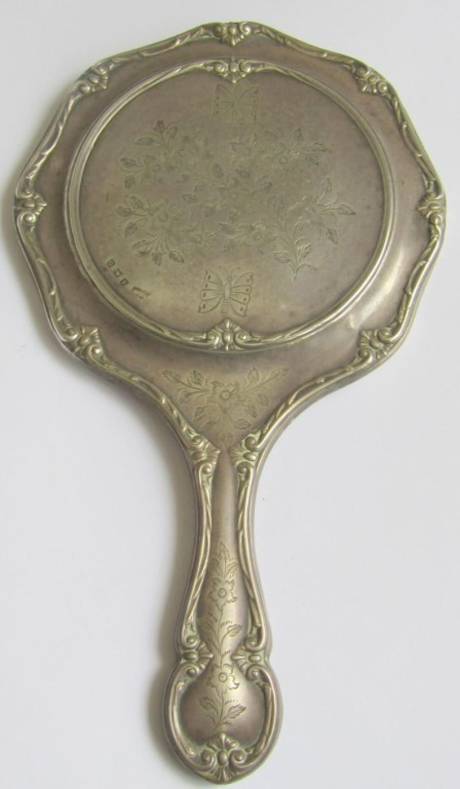 Ornate hand mirror with an oval reflecting surface and engraved floral and butterfly-like designs in a decorative frame.