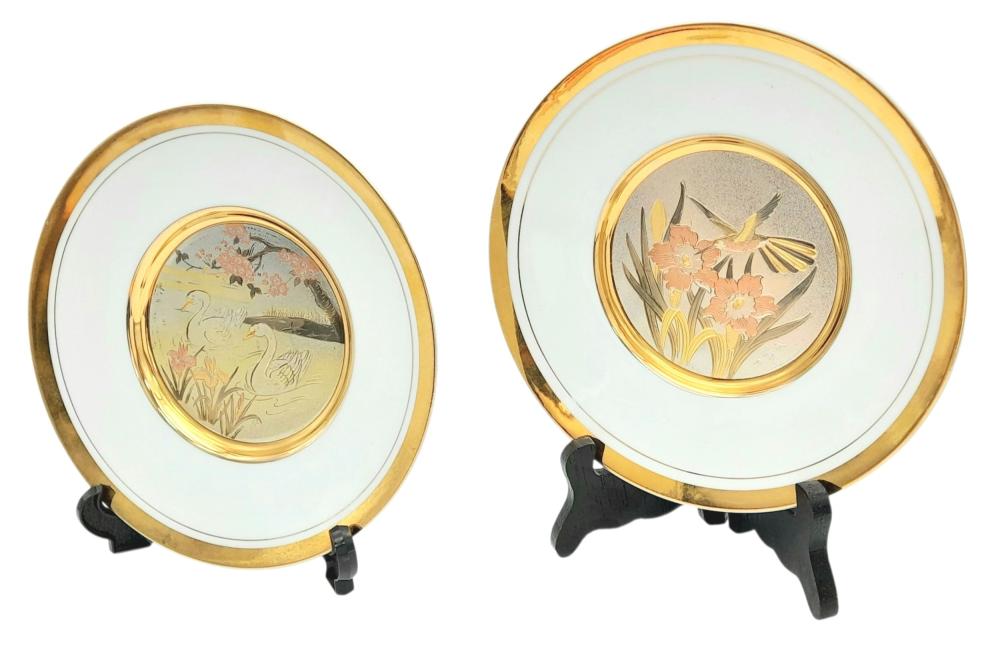 Two round decorative plates with gold rims on black display stands, each featuring a central floral bird motif in soft colors.