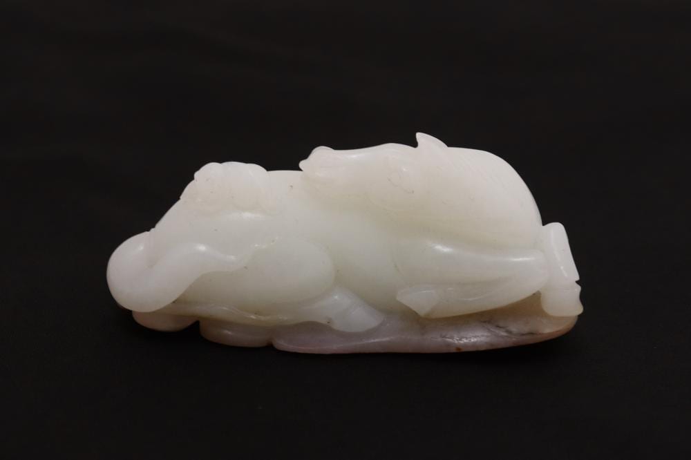 Carved pale jade-like sculpture of a reclining animal with detailed legs and head on a dark background.