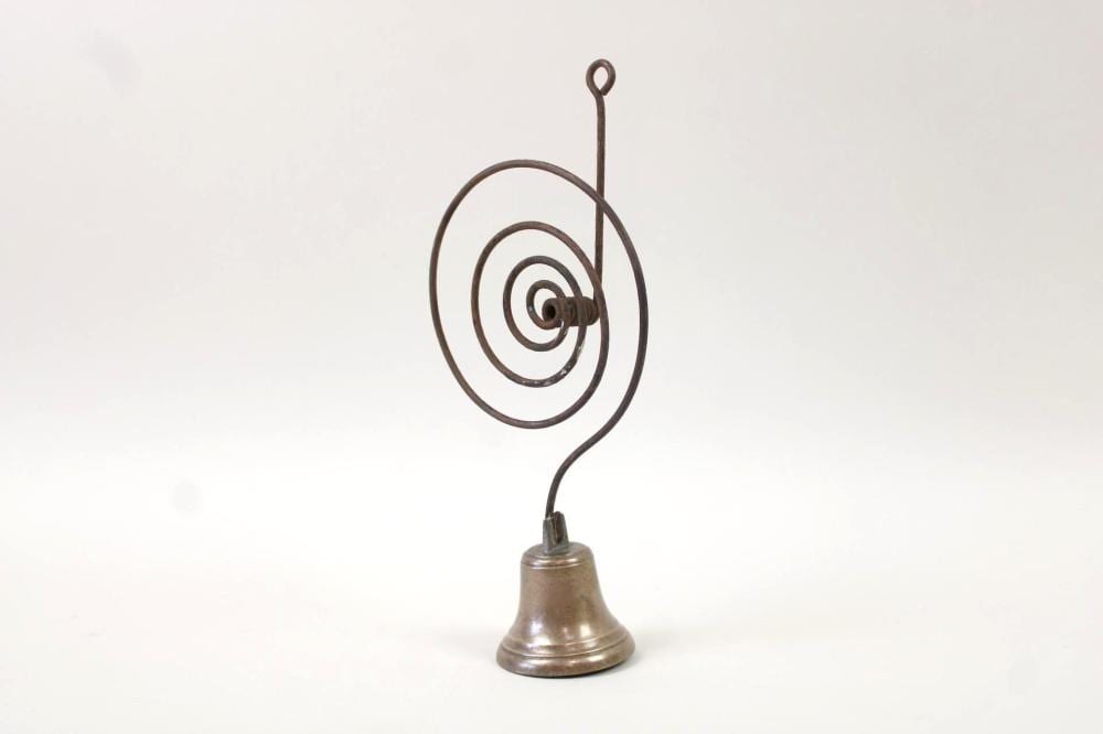 Small metal hand bell with a spiral wire loop on a stand, shown against a plain light background.