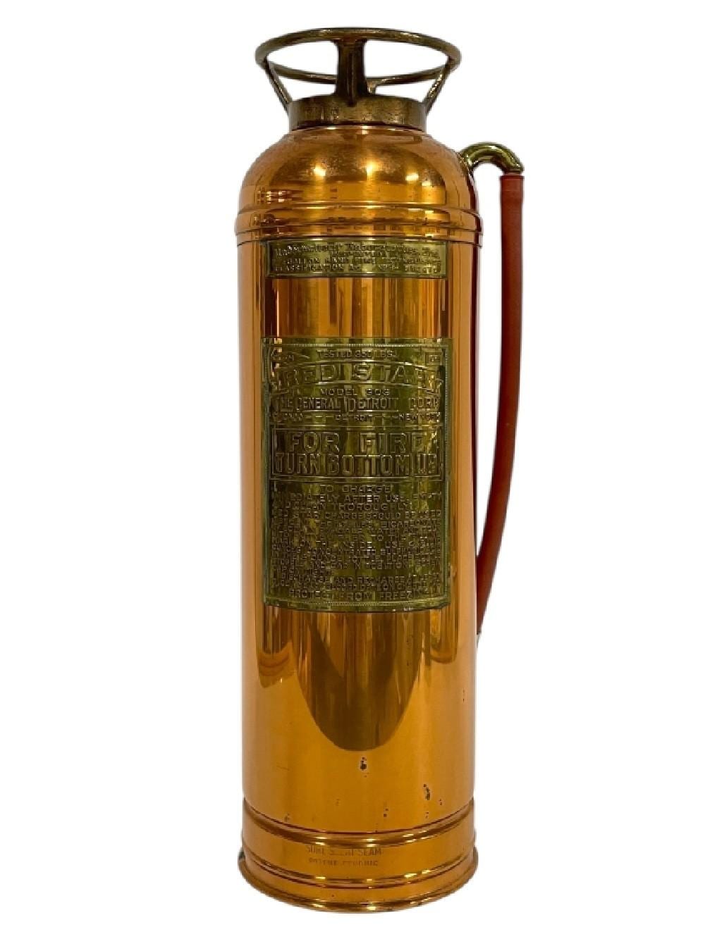 Brass-colored copper fire extinguisher with a top handle and engraved label, photographed against a white background.