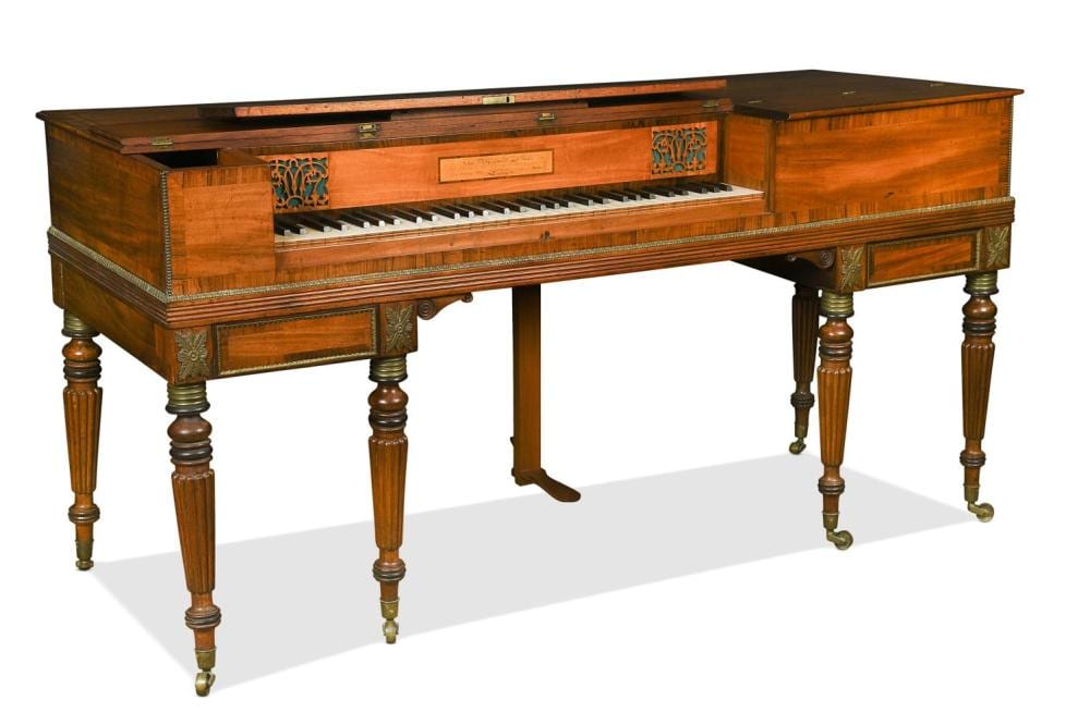 Wooden square upright piano with open keyboard and decorative side panels, shown on four turned legs with brass tips.
