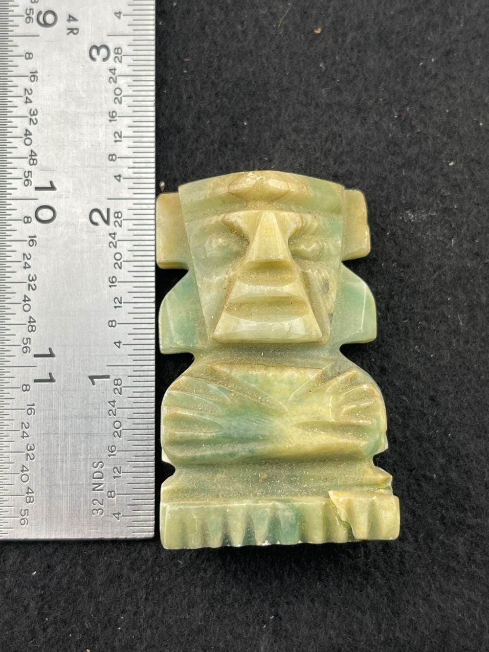 Carved green stone figure with a stylized face and headdress, photographed next to a ruler for scale.