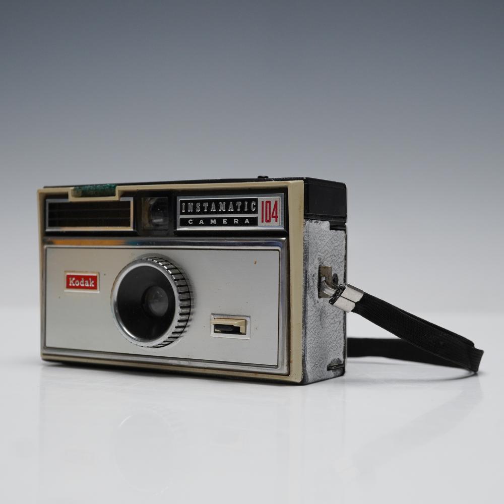 Front view of a Kodak Instamatic 104 vintage film camera with a black lens and viewfinder window, shown on a light background.