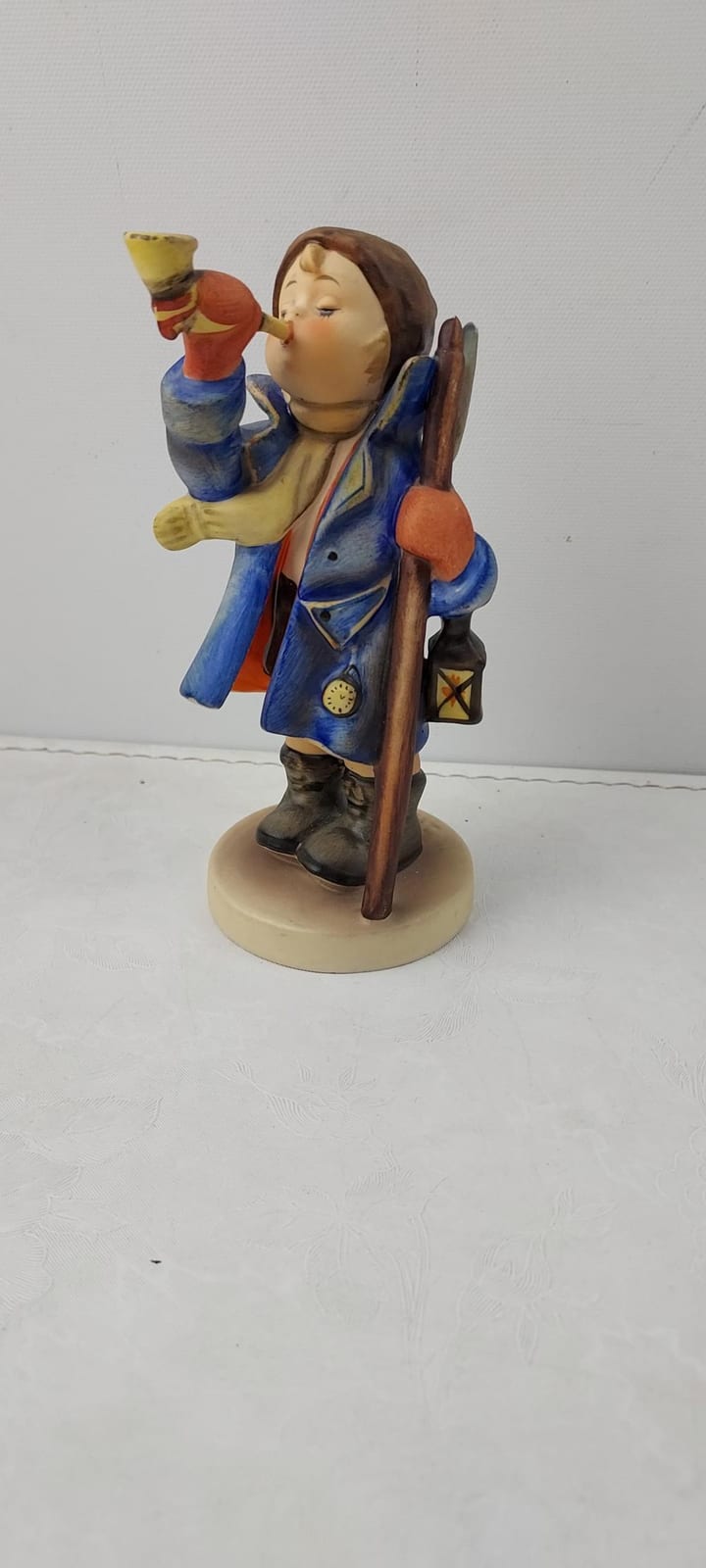 Colorful ceramic figurine of a small child dressed in a blue coat, holding a lantern and waving while walking on a round base.