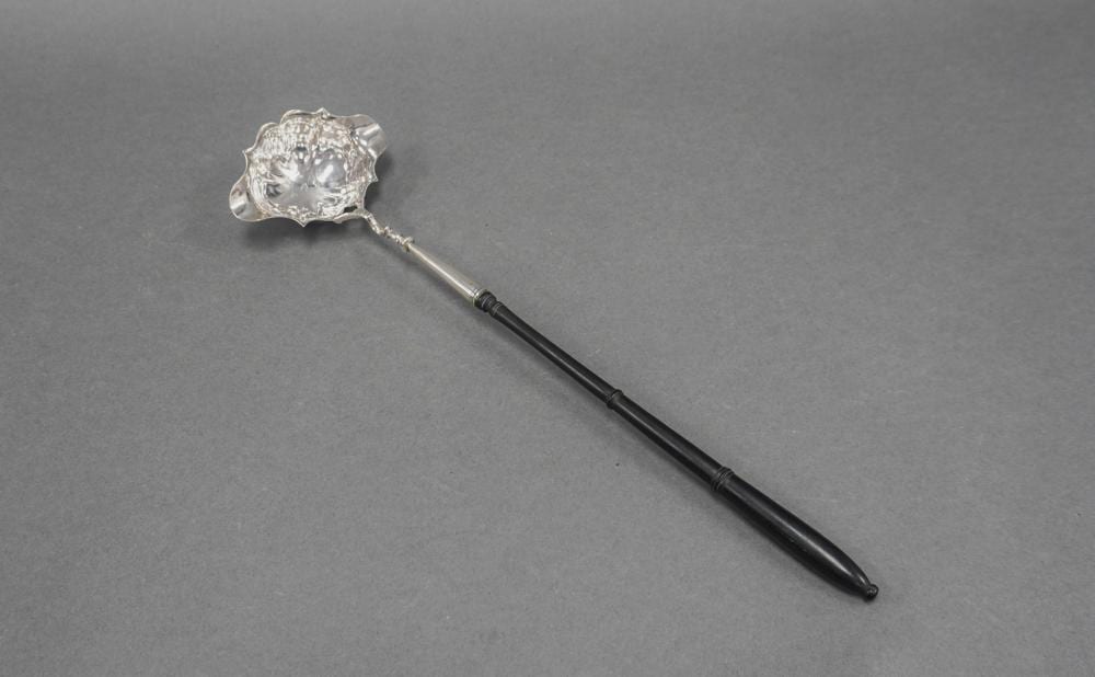 Silver metal ladle with an ornate scalloped bowl and a long dark handle on a gray background.