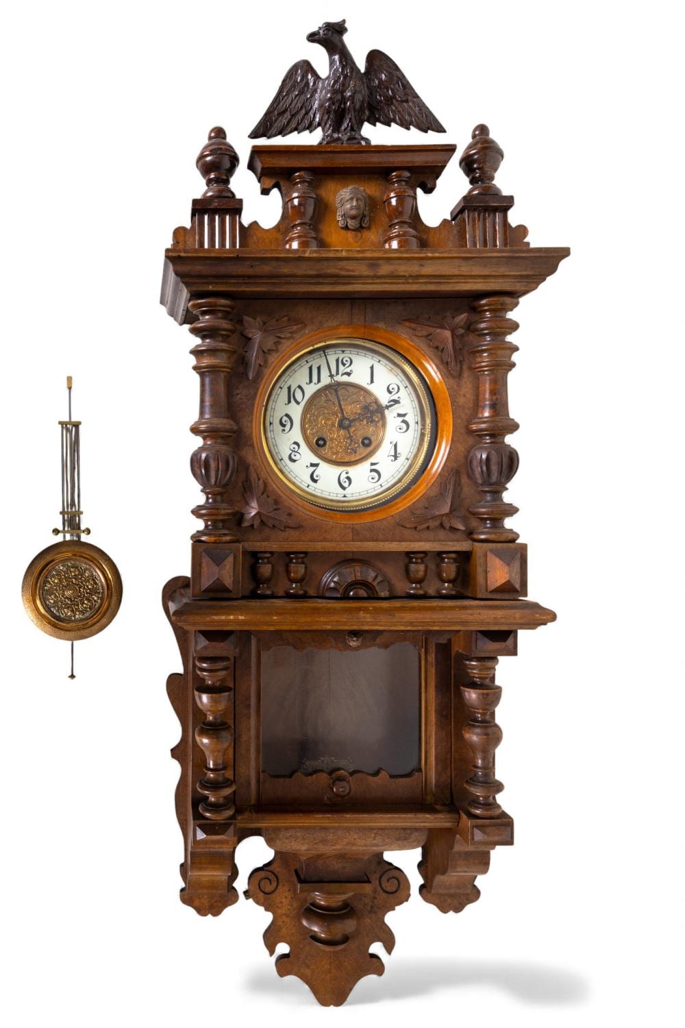 Wooden wall clock with carved case, eagle finial on top, and a round pendulum-less clock face with Arabic numerals.
