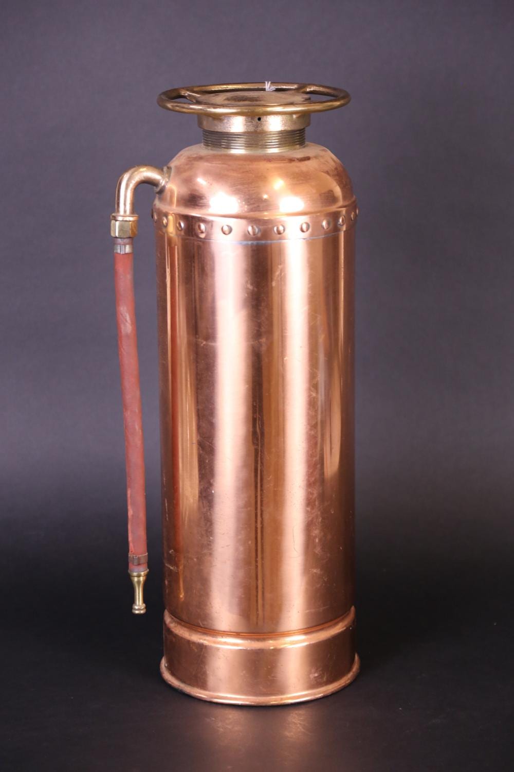 Vintage brass and copper fire extinguisher with a rounded top, hose, and valve, shown against a dark background.