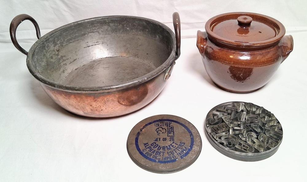 Two copper-colored pots on a white cloth: a large open bowl pot with side handles, and a lidded small pot; a metal baking mold and a round tin are on the table.