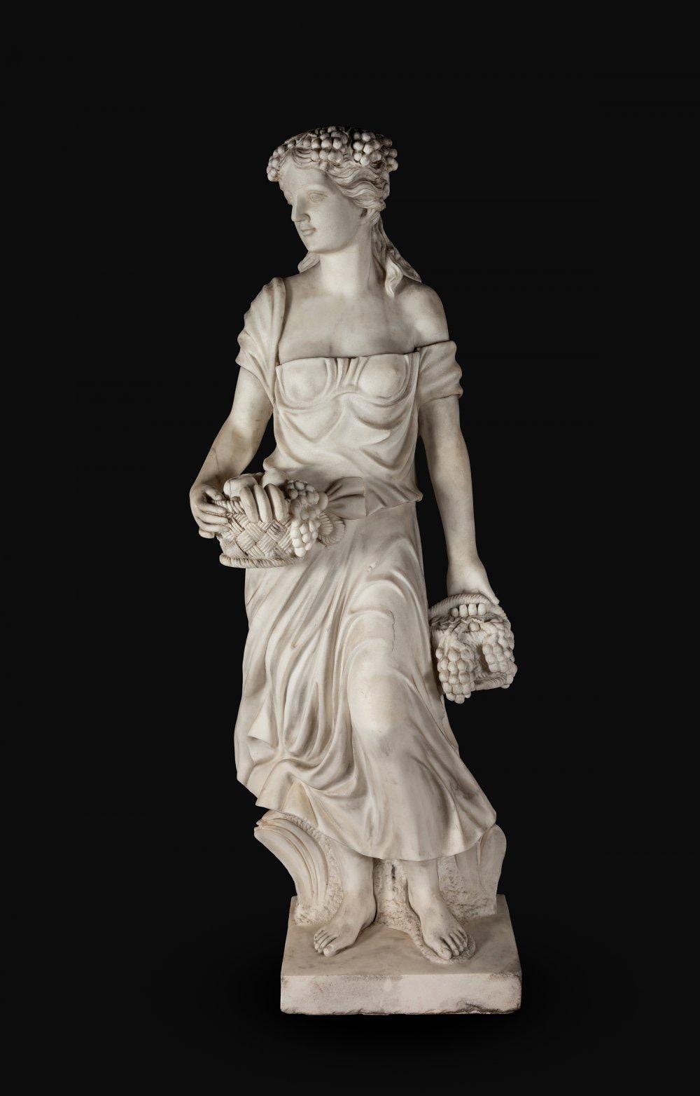 Marble sculpture of a robed female figure standing in profile, holding a bunch of grapes and a basket-like bundle.