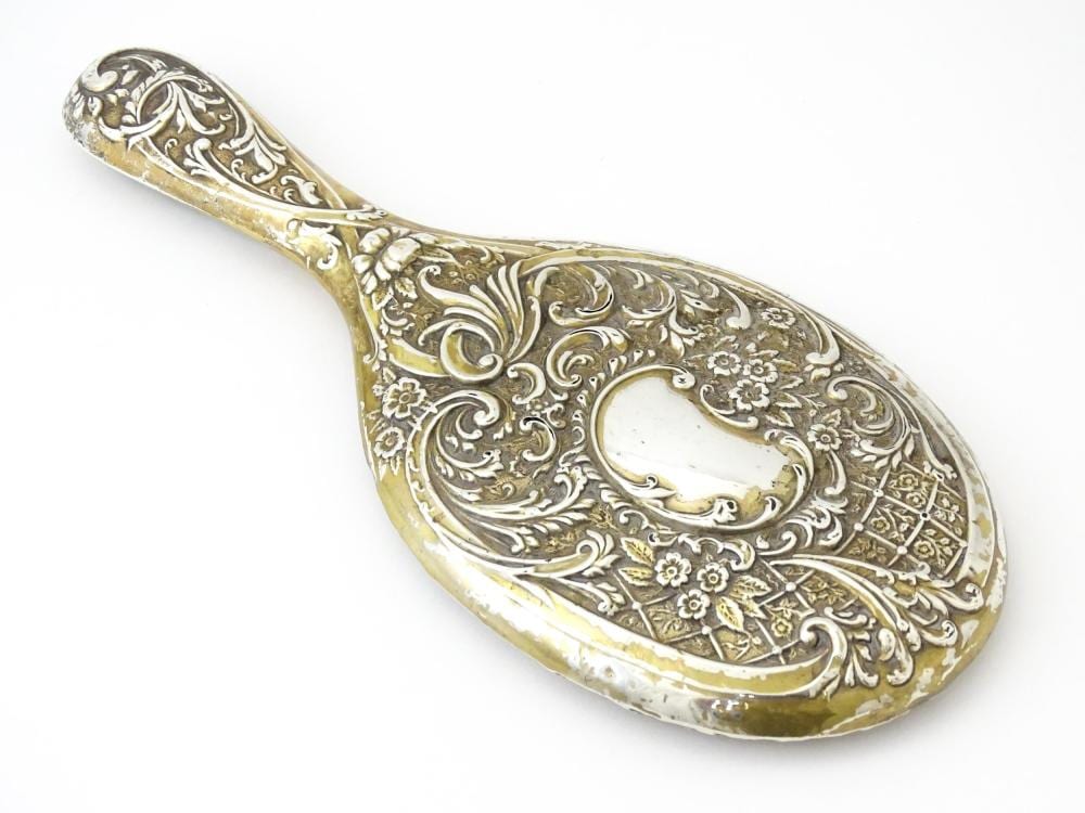 Hand mirror with ornate embossed and engraved floral scrollwork on a silver-colored handle and frame, viewed against a white background.