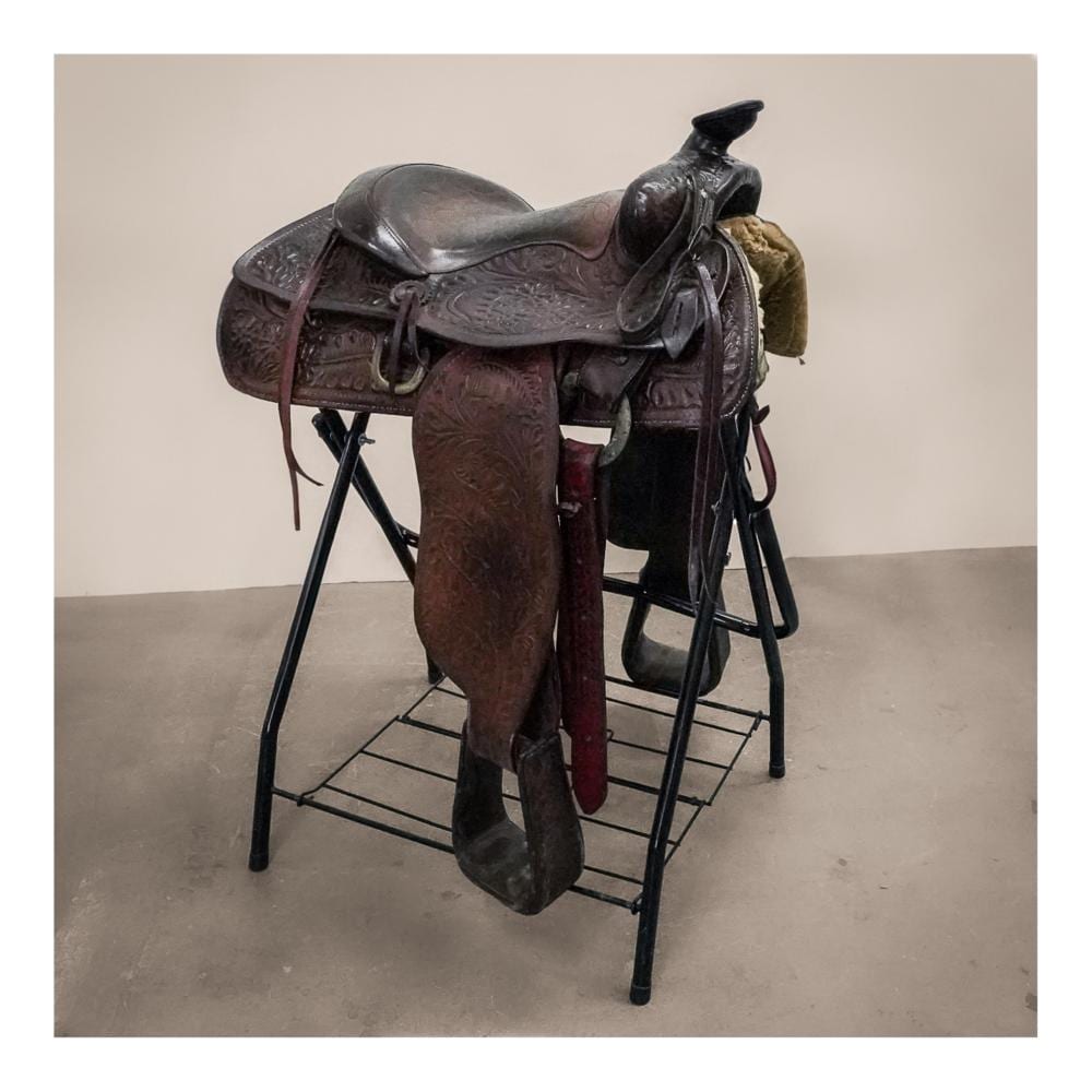 Vintage brown leather Western saddle with dark metal hardware and stirrups mounted on a black display stand.