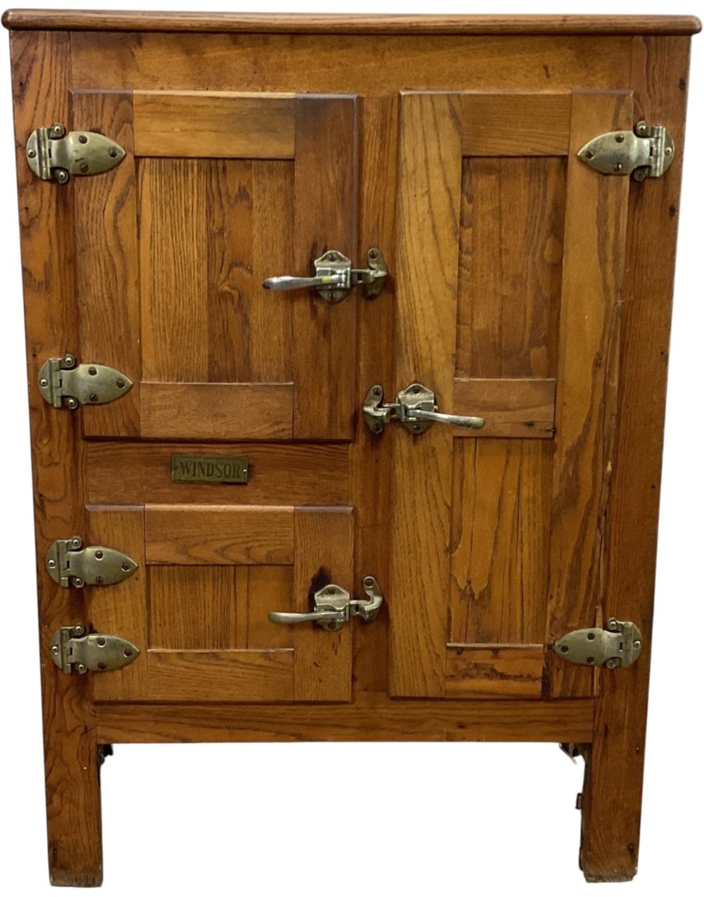 Front view of an antique wooden ice box with a hinged door, metal corner hardware, and a small label reading “WINDSOR.”