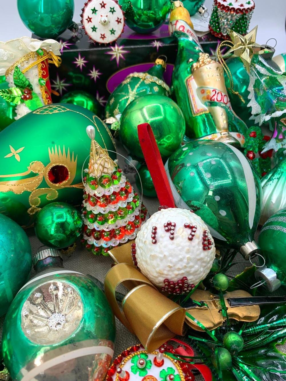 Mixed green Christmas ornaments, including shiny ball ornaments and a beaded tree-shaped ornament, shown piled together with other holiday decorations.