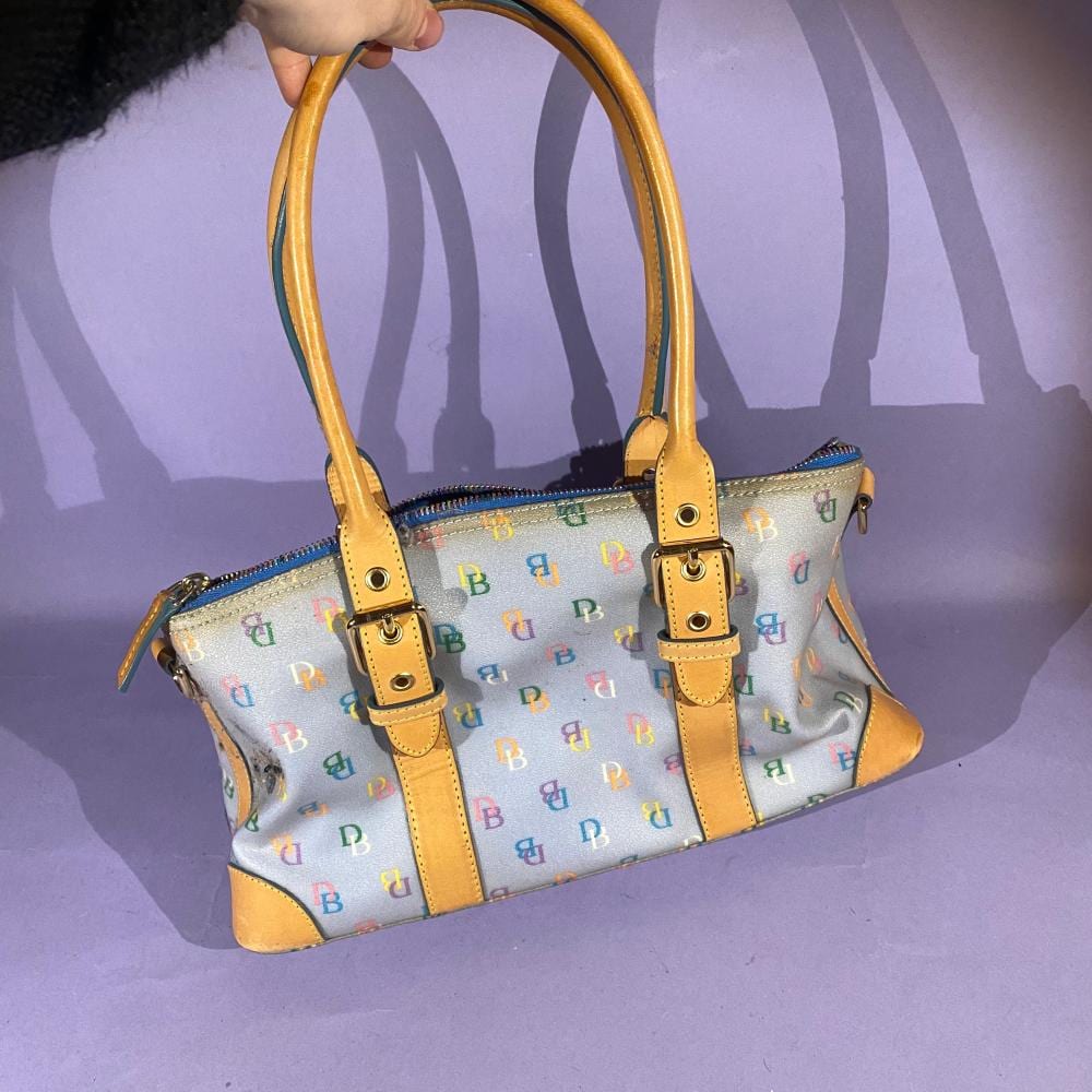 Light blue handbag with tan leather handles and trim, printed with small repeating “B” letters in multicolor, shown on a person’s arm.