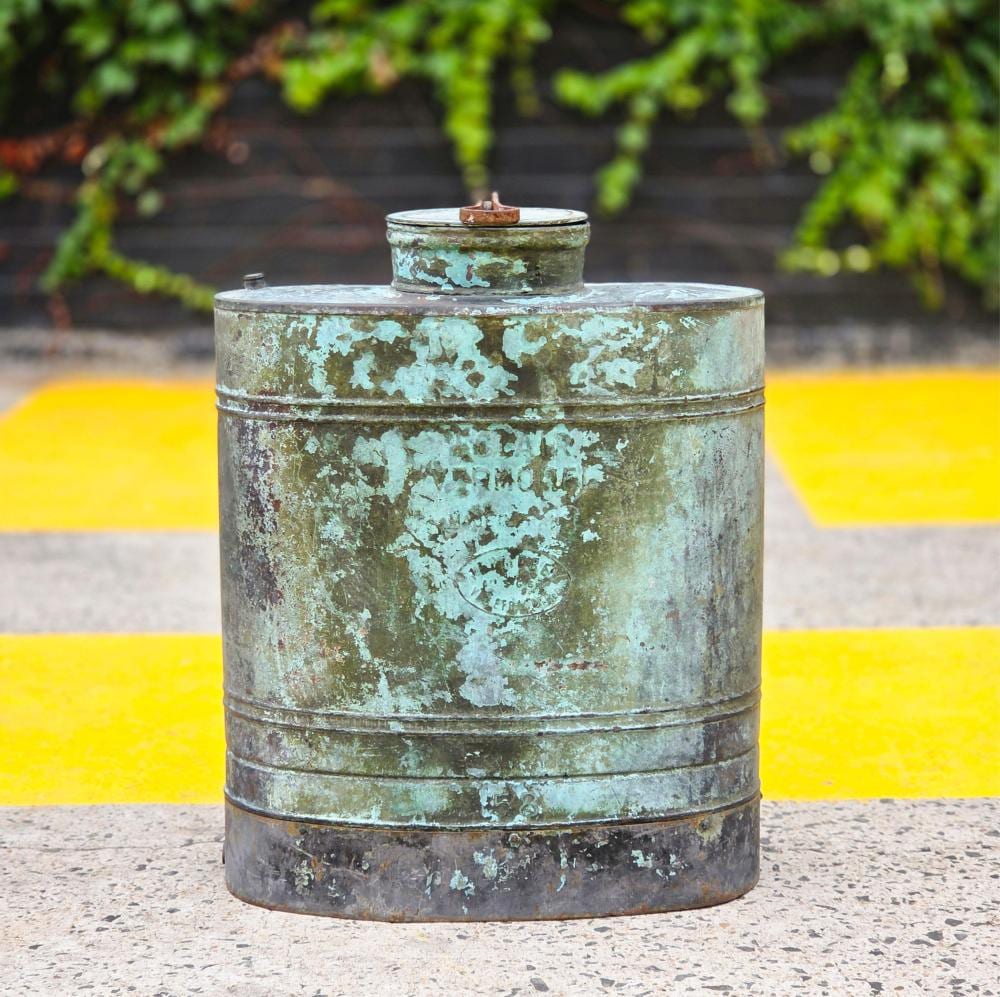 Small cylindrical metal tank with a domed top and a short nozzle, coated in weathered green patina, outdoors on a pavement surface.