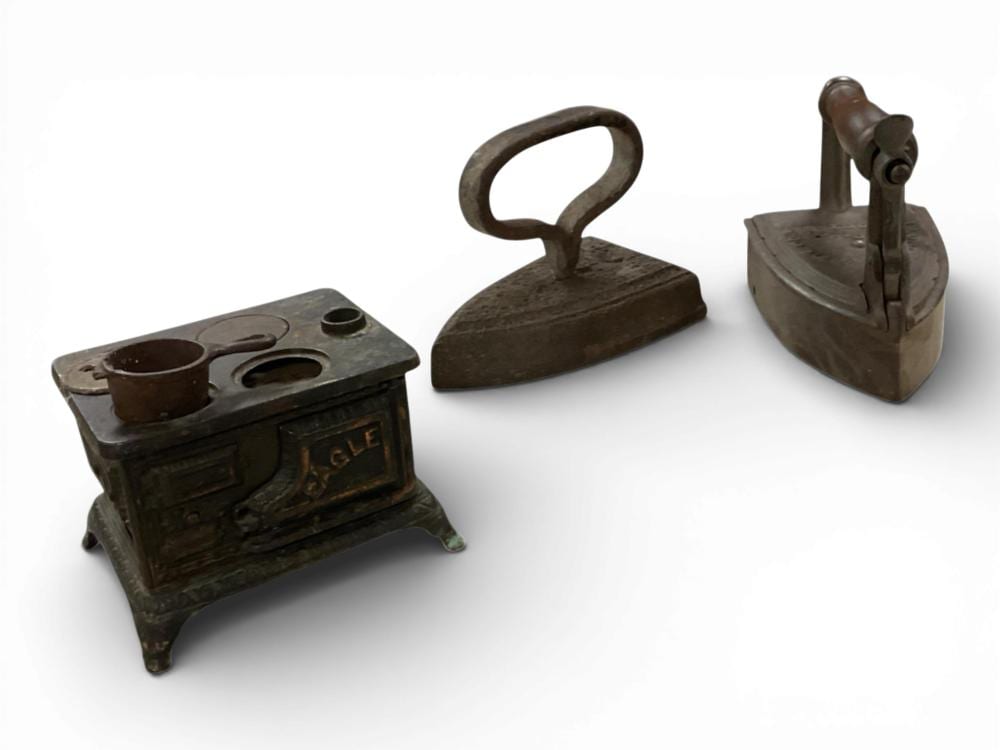 Three small cast iron toy household appliances: a toy stove with two round holes, a flat iron with handle, and another flat iron.