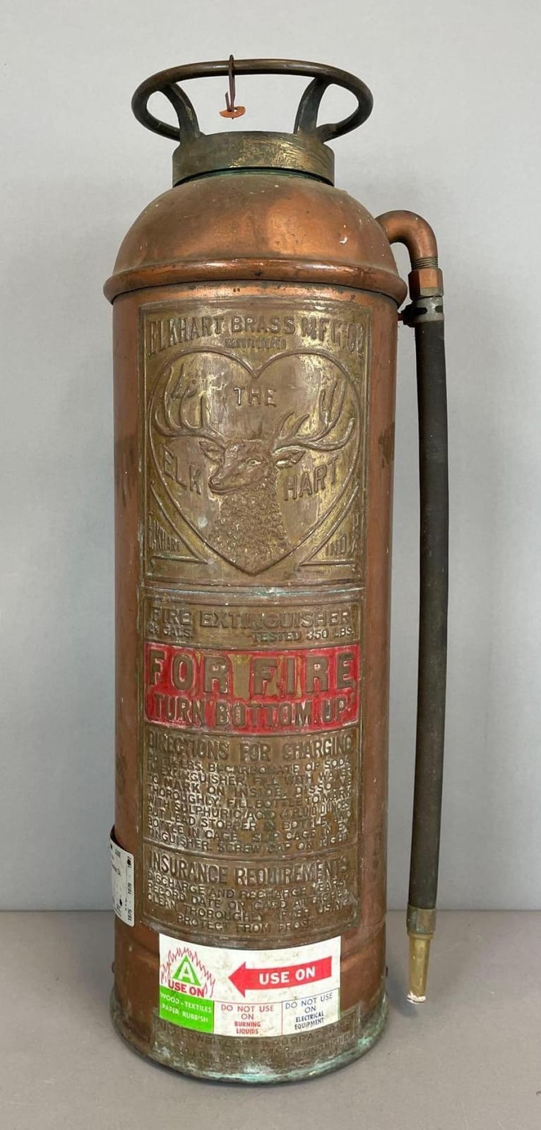 Antique brass fire extinguisher cylinder with carrying handle and hose, featuring embossed badge and red label text.