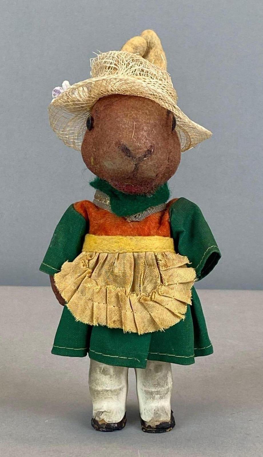 Vintage toy bunny dressed in a green and orange outfit with a straw hat, standing against a plain gray background.