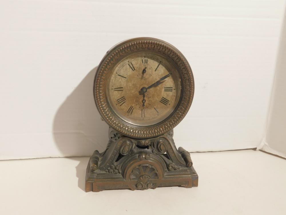 Round face mantel clock with Roman numerals, ornate metal bezel, and decorative base on a white background.