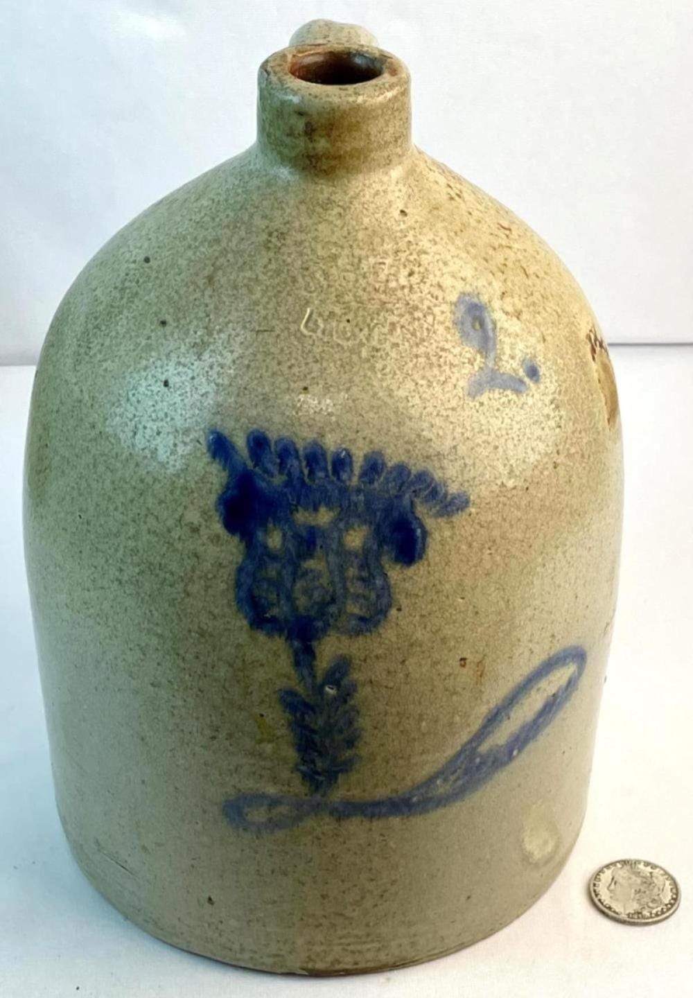 Close-up of a small antique stoneware jug with a rounded body, short neck, and blue floral decoration on a greenish glaze.