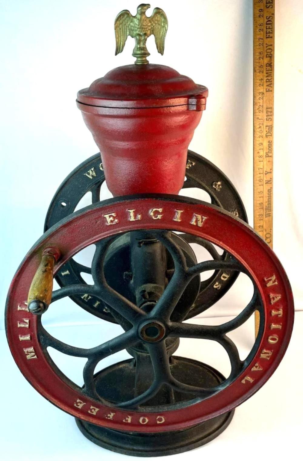 Cast iron countertop coffee grinder with red hopper and eagle finial on top, wheel base, and black crank handle.
