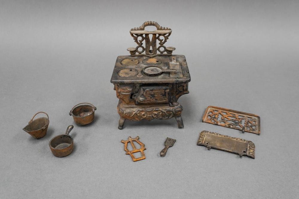 Small cast-iron miniature stove with decorative top and side, shown with miniature tools and accessories laid out nearby.