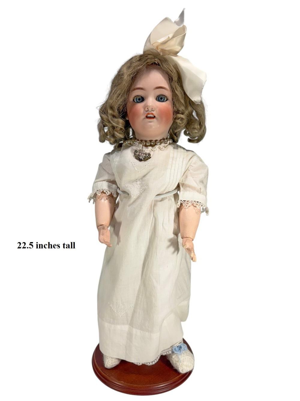Porcelain girl doll with blue eyes and curly brown hair, wearing a white dress with lace trim and a cream bow on her head.