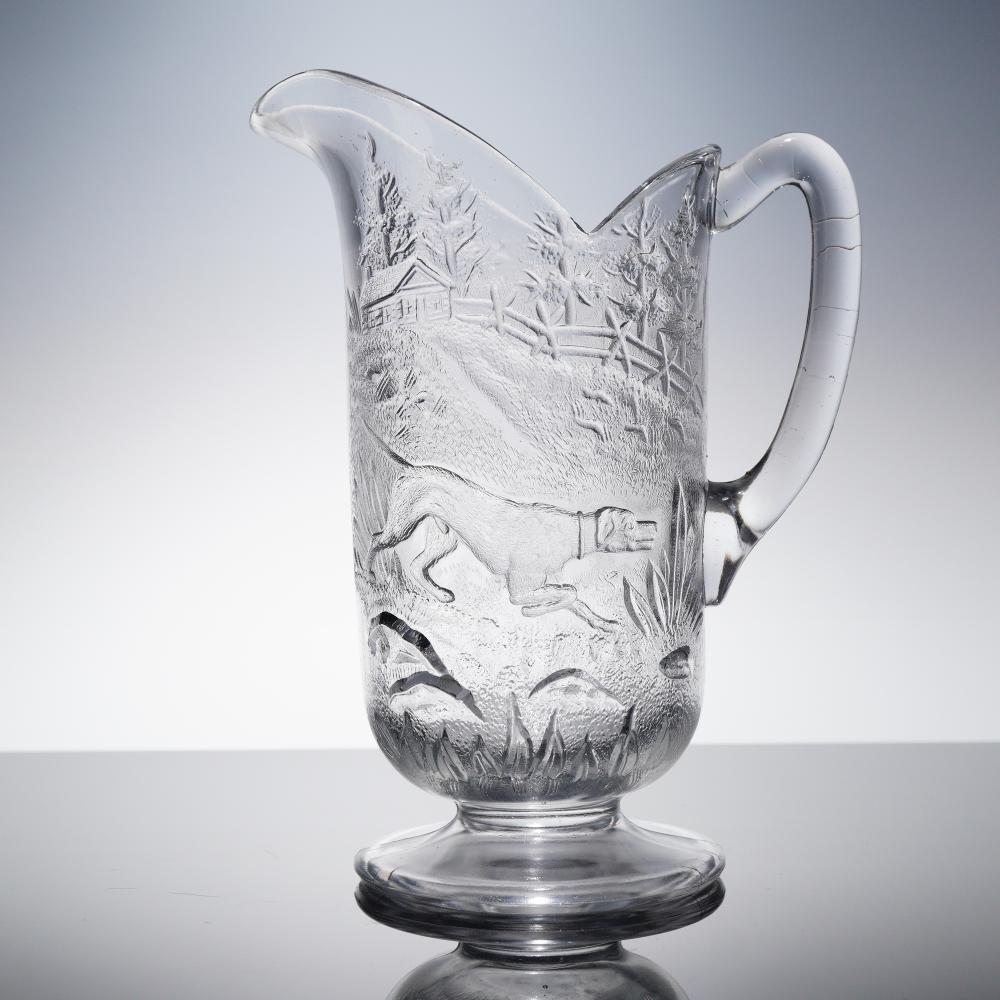 Clear glass water pitcher with a molded relief scene of hunters, dogs, and trees on its body.