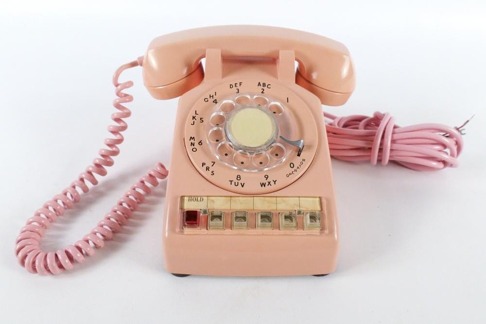 Pink rotary dial desk telephone with coiled cord and lettered number dial on a white background.