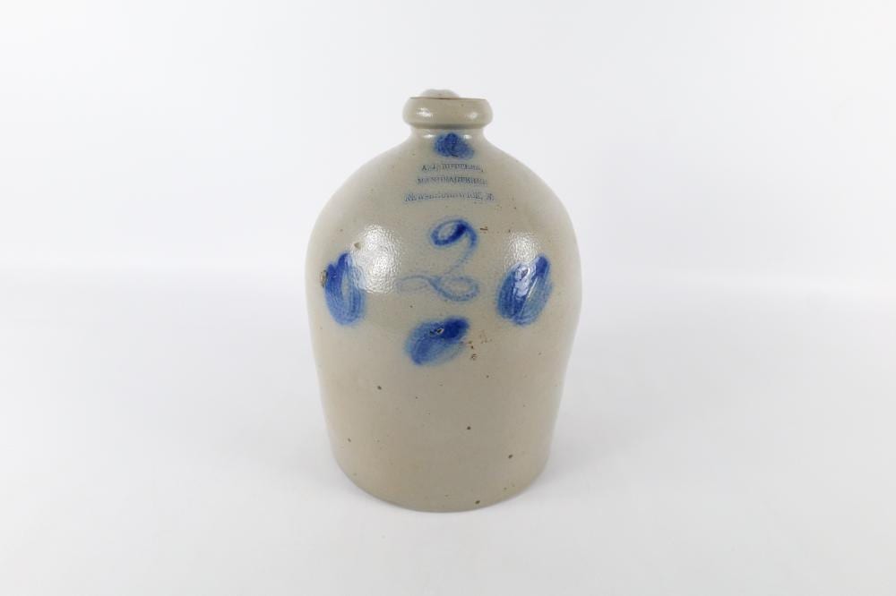 Small cream-colored stoneware jug with a blue painted decorative motif on the front and a narrow neck and stopper-like top.