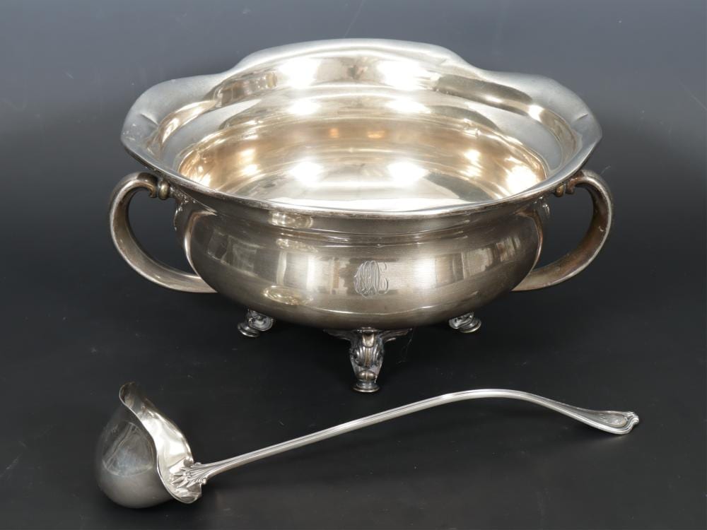 Close-up of a polished silver punch bowl with two side handles on three feet, shown with a long ladle and small ladle base.