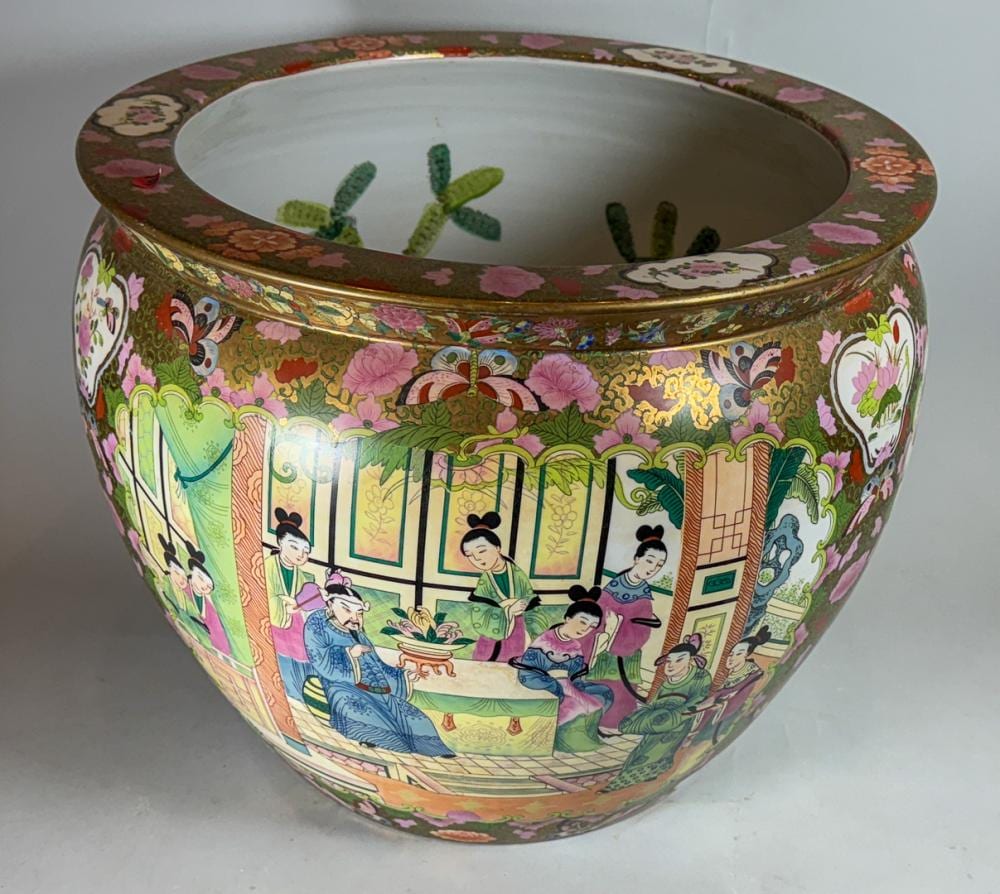 Large Chinese-style fishbowl with ornate floral rim and a painted scene of people in a courtyard, holding green plants inside.