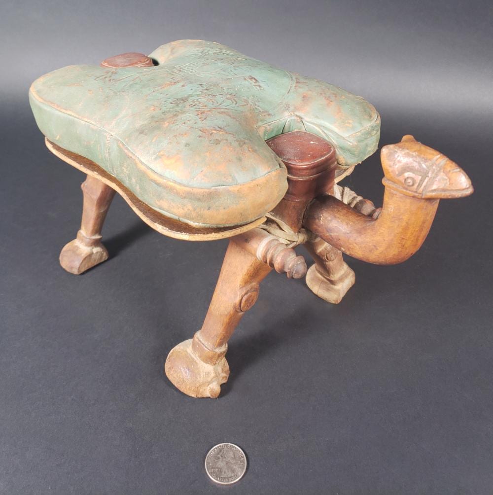 Vintage carved wooden stool shaped like a camel saddle form with a green cushioned top and front legs; small coin shown for scale.